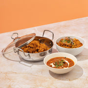 Vinod stainless steel kadai with glass lid containing aromatic biryani rice dish alongside Indian curry and rice bowls on marble surface with orange background